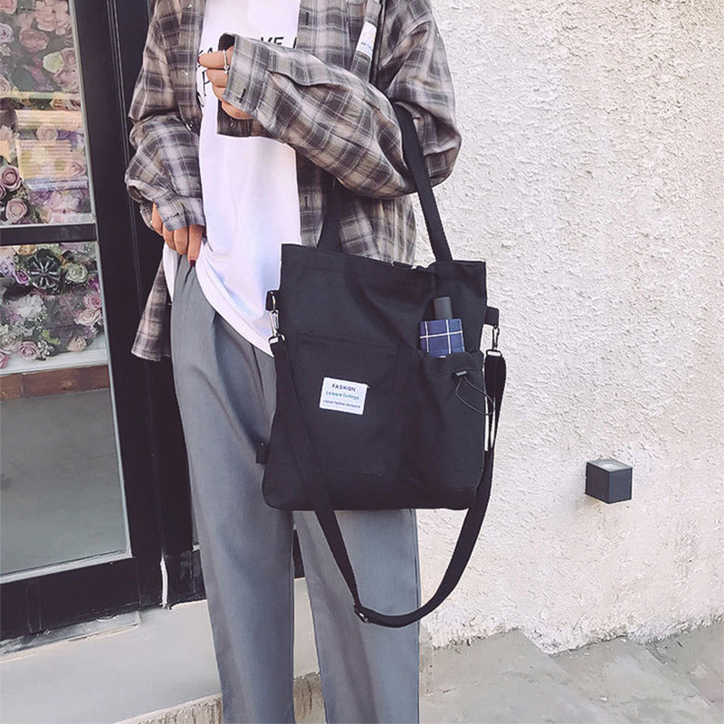 Black canvas tote bag with front pockets, plaid patch sleeve, and adjustable strap